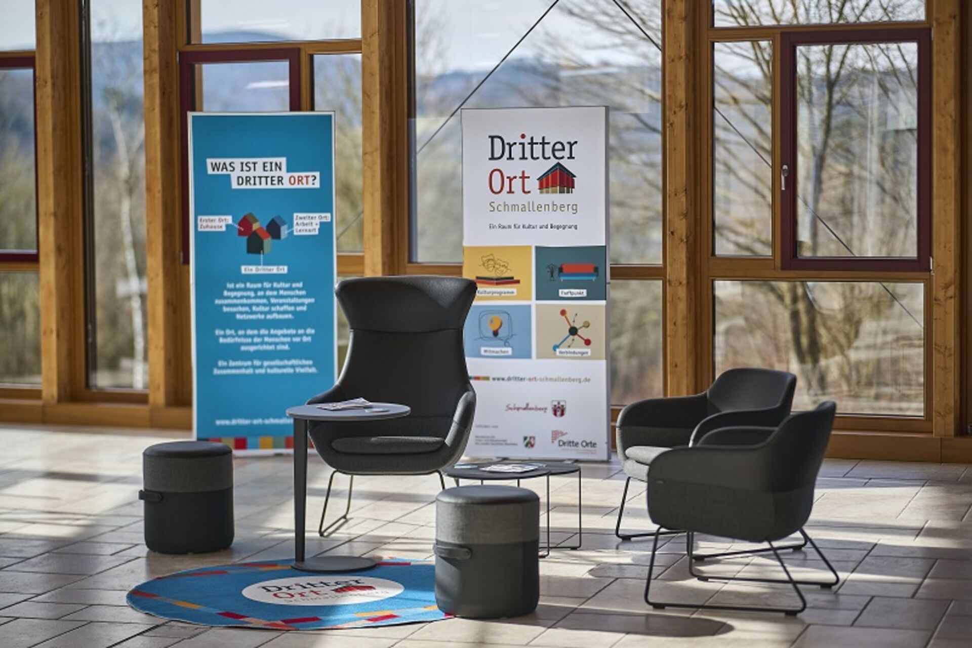 Der Dritte Ort Schmallenberg im Holz- und Touristikzentrum wurde als neuer Ort für Kultur und Begegnung im Mai 2025 eröffnet. Das Bild zeigt Sessel und Hocker vor zwei Leuchtwerbungen im Saal des Holz- und Touristikzentrums.